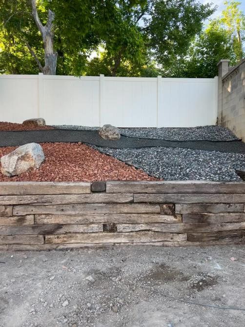 A tiered garden bed with red and gray gravel, large rocks, and a wooden retaining wall in front of a white fence.