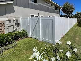 White vinyl fence with a gate, bordering a grassy yard next to a two-story house with flowers in the foreground.