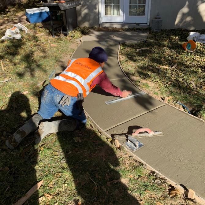 A worker wearing an orange safety vest kneels on a new concrete sidewalk, smoothing the surface with a trowel outdoors.