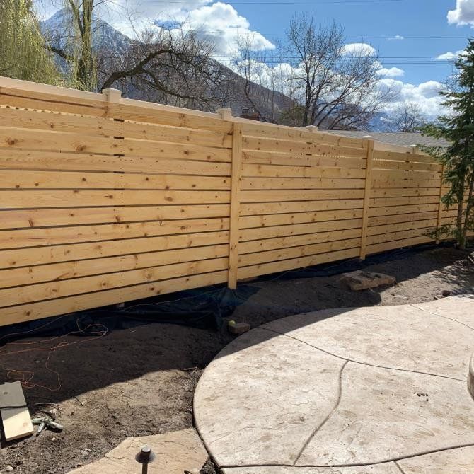 Wooden horizontal slat fence in a yard with a concrete patio, trees, and mountains in the background.