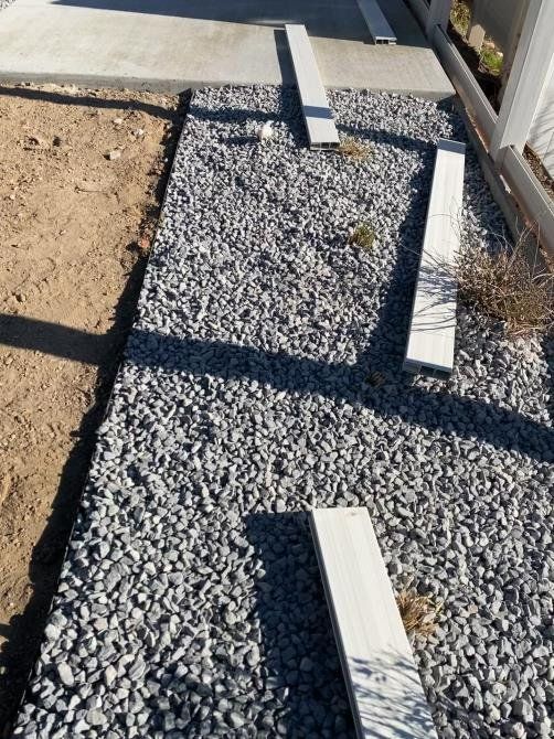 Gravel path with gray stepping stones leading to a concrete walkway.  The side has dirt and a white fence.
