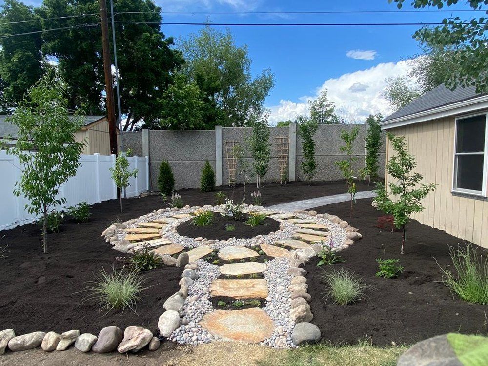 Stone pathway leading to a circular garden bed in a yard with trees, a fence, and a small building.