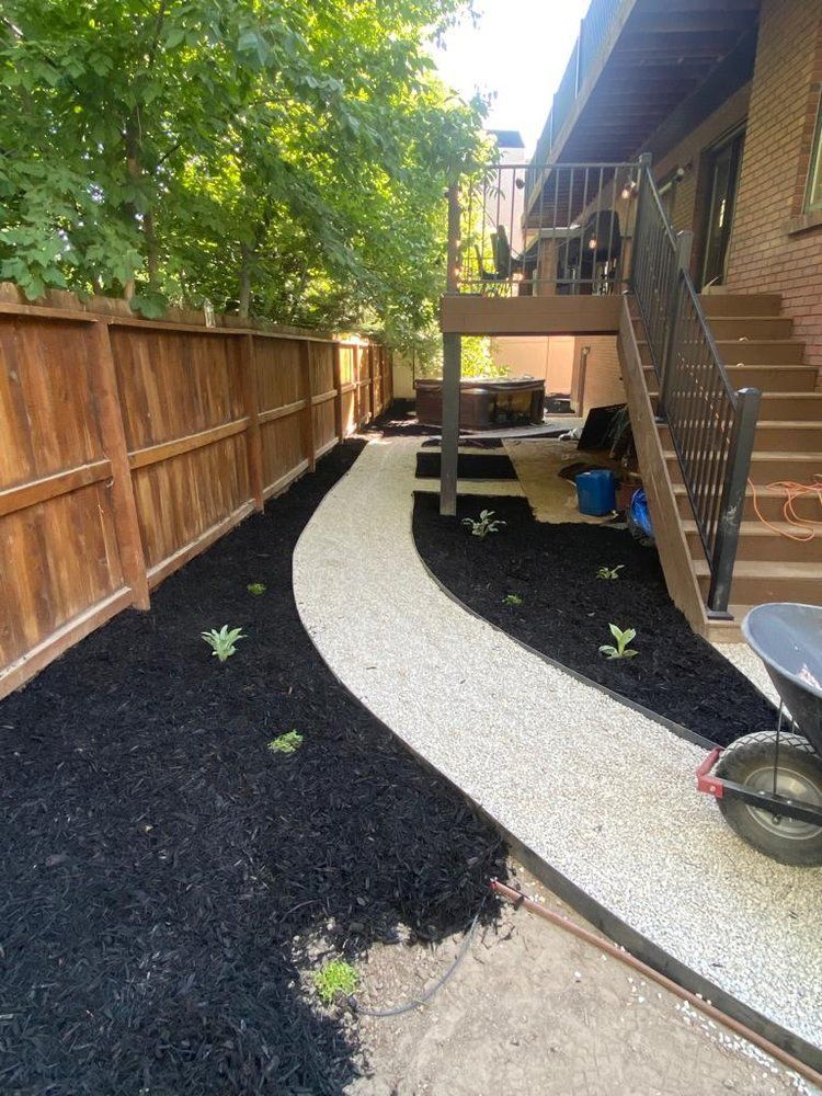 Gravel path winds through a landscaped backyard with black mulch and a wooden fence. Steps lead up to a deck.
