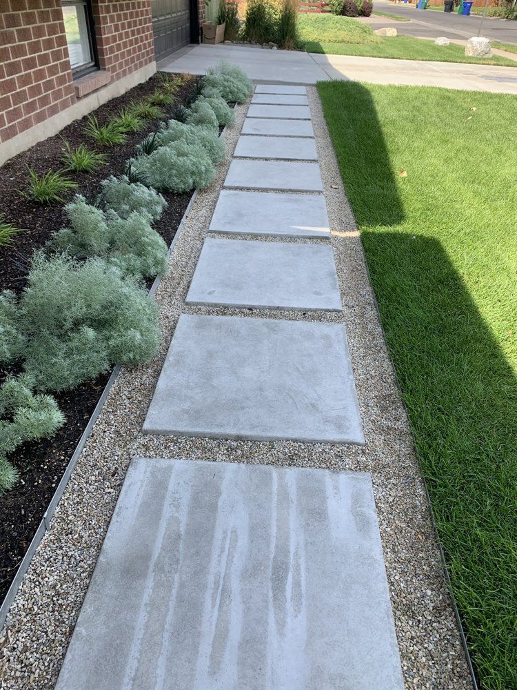 A paved walkway with large, gray square stepping stones, gravel, and plants, leading toward a brick building and lush green lawn.