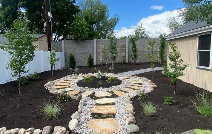 A stone pathway leads through a backyard garden with trees, flowers, and a shed, under a blue sky.