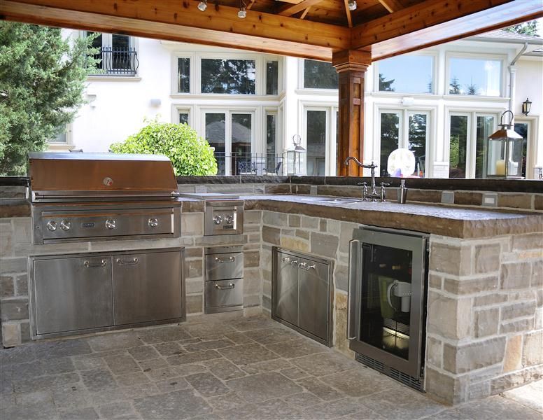 Outdoor kitchen with stainless steel appliances built into a stone structure, under a wooden pergola, near a large house with many windows.