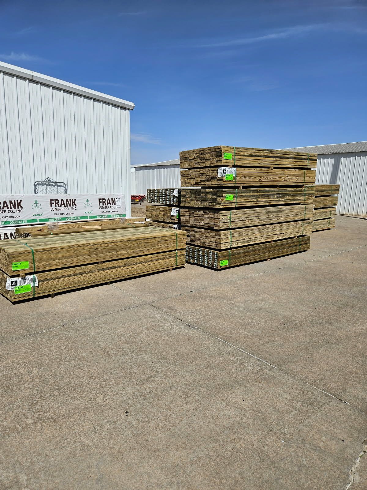 Piles of treated lumber stacked outside under a clear, blue sky. Large white building in the background.