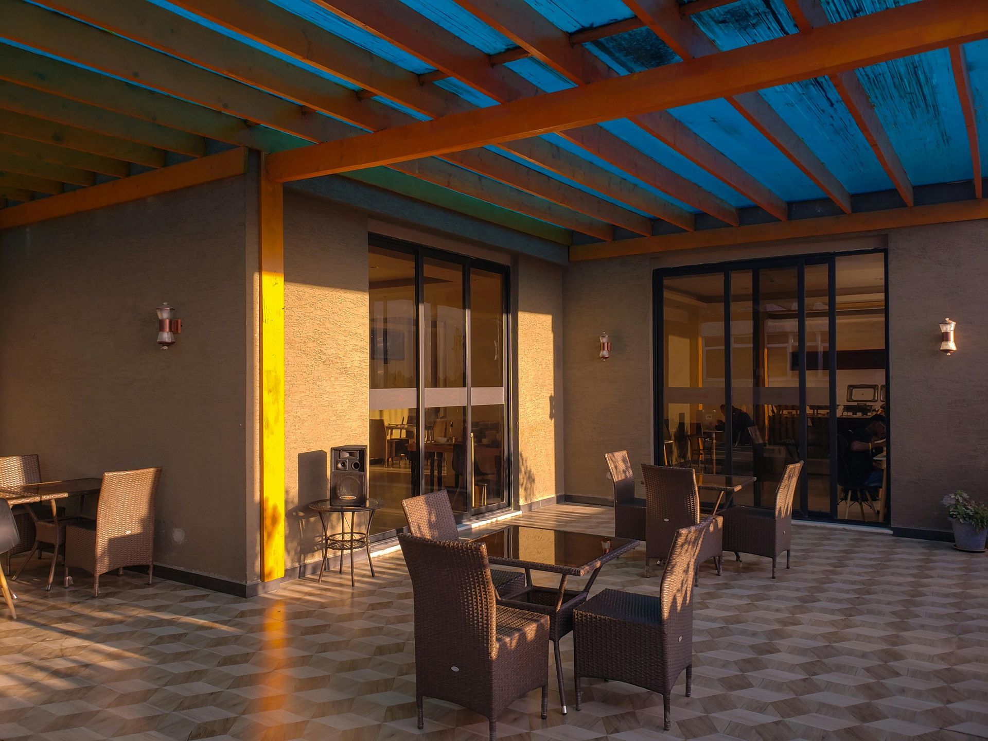 Outdoor patio with tables and chairs under a wooden pergola with blue roofing. Evening light casts shadows.