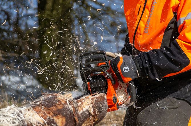 Worker in orange safety gear using a chainsaw on a log with flying woodchips.