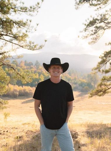 Man in cowboy hat smiles, standing outdoors with a mountain backdrop.