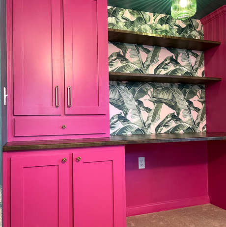 Bright pink built-in desk with cabinets, shelves, and tropical leaf wallpaper.