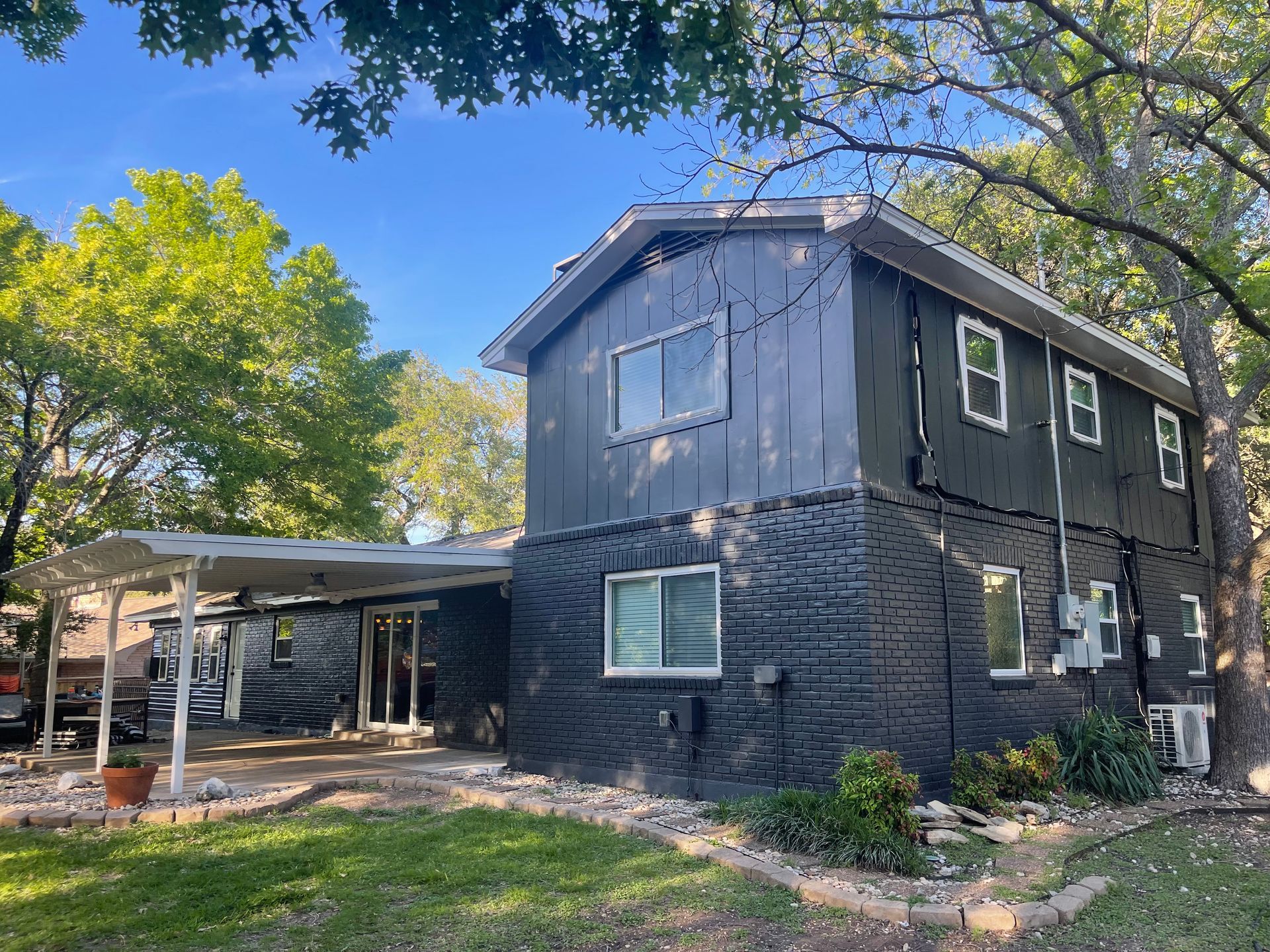 Two-story house with black painted siding and brick exterior. White awning and large trees in the yard.