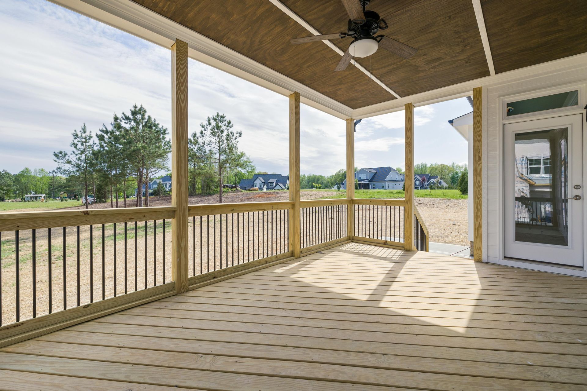 A wooden porch with a ceiling fan, railings, and a glass door overlooking a grassy backyard and distant houses.