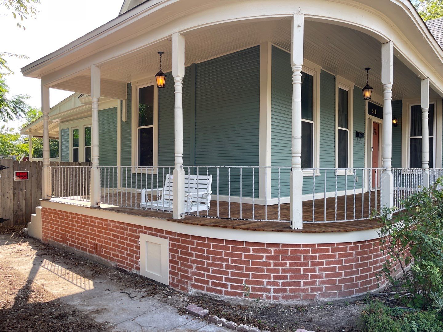 A teal house with a wraparound porch, brick foundation, and white railing.