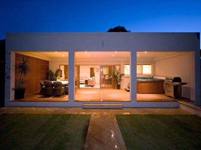 An illuminated patio at night, featuring a dining area, open living space, and hot tub under a flat-roofed structure.