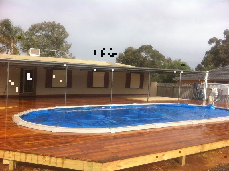 An oval swimming pool with a blue cover, set into a wooden deck outside a house with a covered patio.