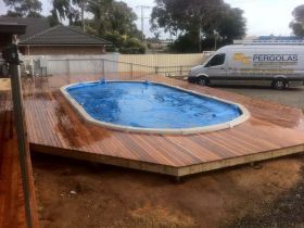 An oval above-ground pool covered with a blue thermal blanket, surrounded by a wooden deck in a yard under construction.