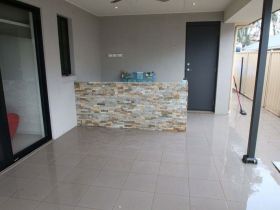 A covered patio with light-colored tile flooring, a stone-veneer accent wall, and a dark grey door.
