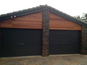 A double garage with dark roller doors, brick pillars, and wooden siding under a dark tiled gable roof.