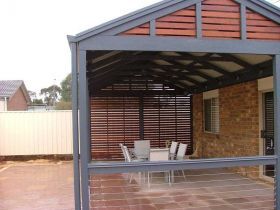 A covered patio with a dark grey frame, wooden slat walls, and a dining set on a paved floor outside a brick house.