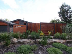 Modern home exterior with a horizontal wooden fence, stone landscaping, and the address number 26 visible on the wall.
