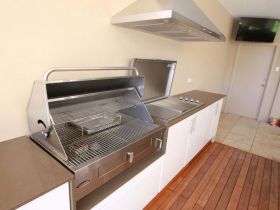 Stainless steel outdoor grill and stovetop setup on a white counter with a range hood on a wooden deck.