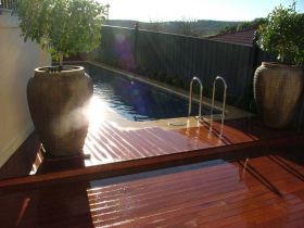 A swimming pool with metallic ladders sits beside a wooden deck and two large stone planters holding small green trees.