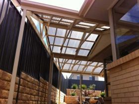 A covered patio with a translucent corrugated roof, beige brick walls, and a black metal fence in a residential backyard.