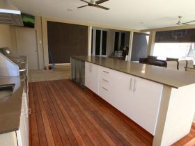 Modern outdoor kitchen with white cabinets, stone countertops, and wooden flooring on a covered patio.