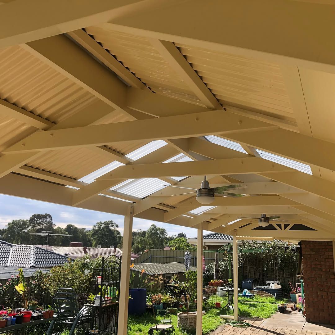 A brown wooden lattice pergola roof with a translucent corrugated covering and two ceiling fans against a brick wall.