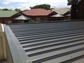 A close-up of a gray corrugated metal roof, with residential houses visible in the background under a cloudy sky.