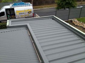A high-angle view of a gray metal roof with a box gutter, with a work van parked on the street in the background.