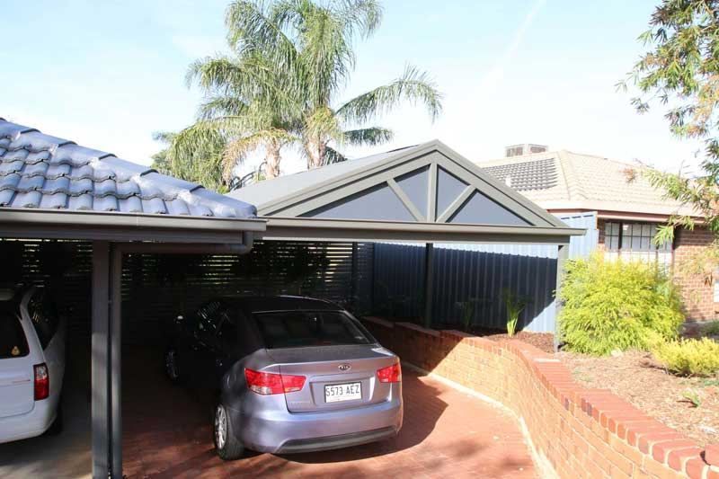 A gray sedan parked under a covered carport next to a suburban house with a brick wall and green landscaping.