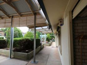 A paved walkway beside a brick wall, leading to a garden area covered by a corrugated metal patio roof.