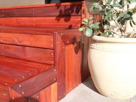 A set of reddish-brown wooden steps with visible screws, positioned next to a large, light-colored ceramic planter.