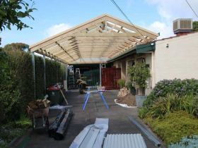 A wooden carport frame under construction attached to a house, with building materials scattered on the concrete patio.
