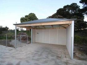 An open-fronted carport with a grey corrugated roof, wooden framing, and light-colored walls on a concrete pad.