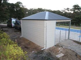 A white outdoor structure with a gray metal roof located next to a swimming pool in a grassy yard.