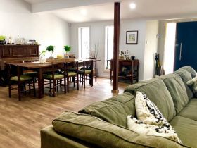 A bright room featuring a wooden dining set, a piano, and a large green sofa, illuminated by natural light from windows.