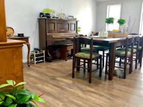 Dining room with a wood table, chairs, and a piano against a light grey wall over a wood-look floor.