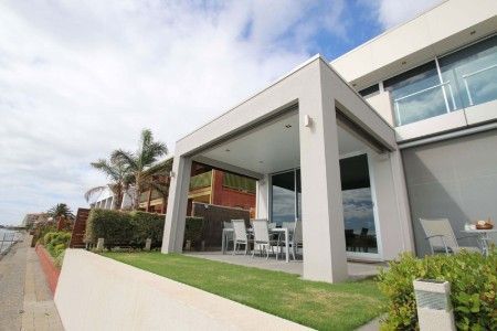 A modern patio with white columns and outdoor furniture overlooks a grassy yard and nearby water under a cloudy sky.