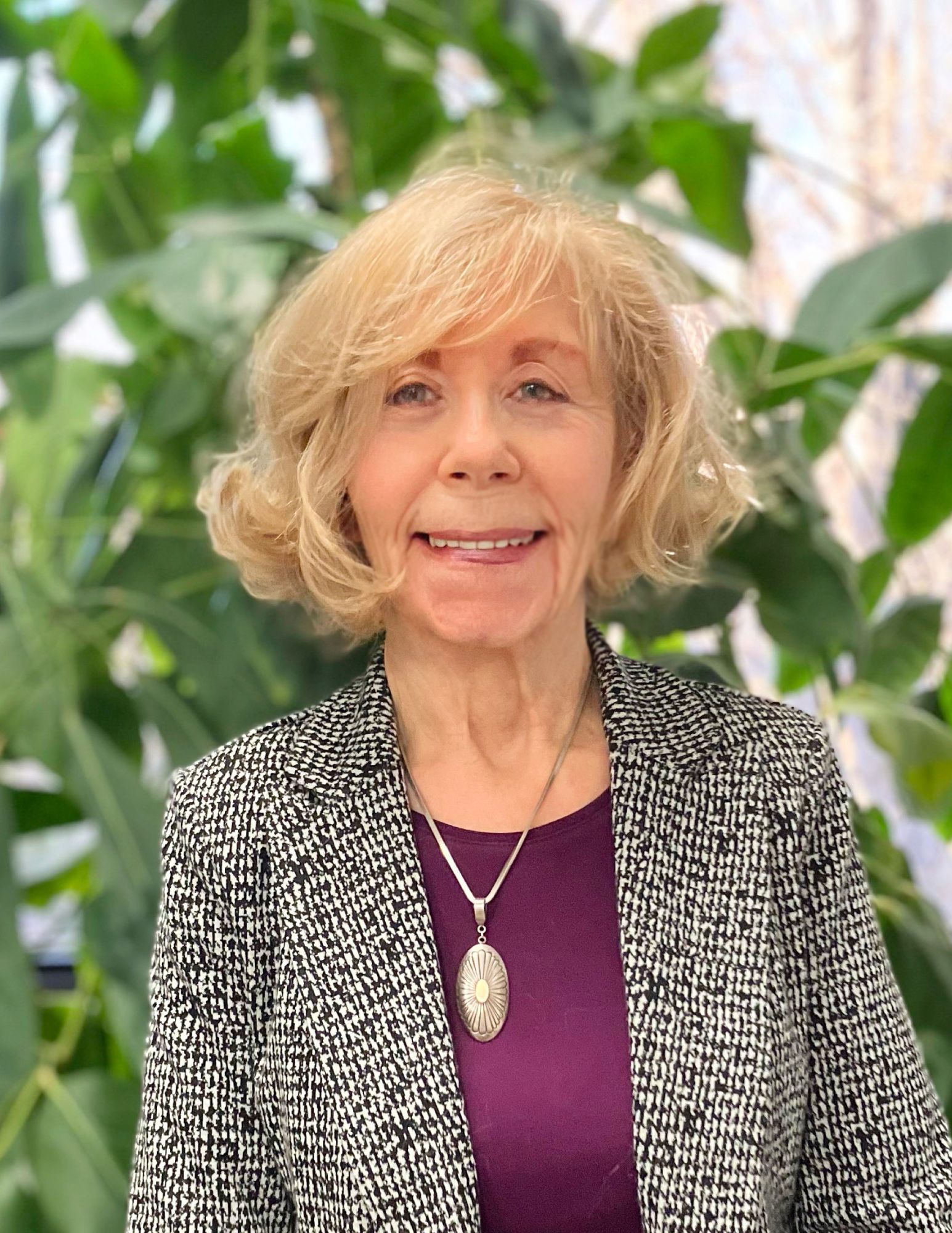 Woman with blonde hair wearing a patterned jacket and purple shirt smiles, plants in the background.