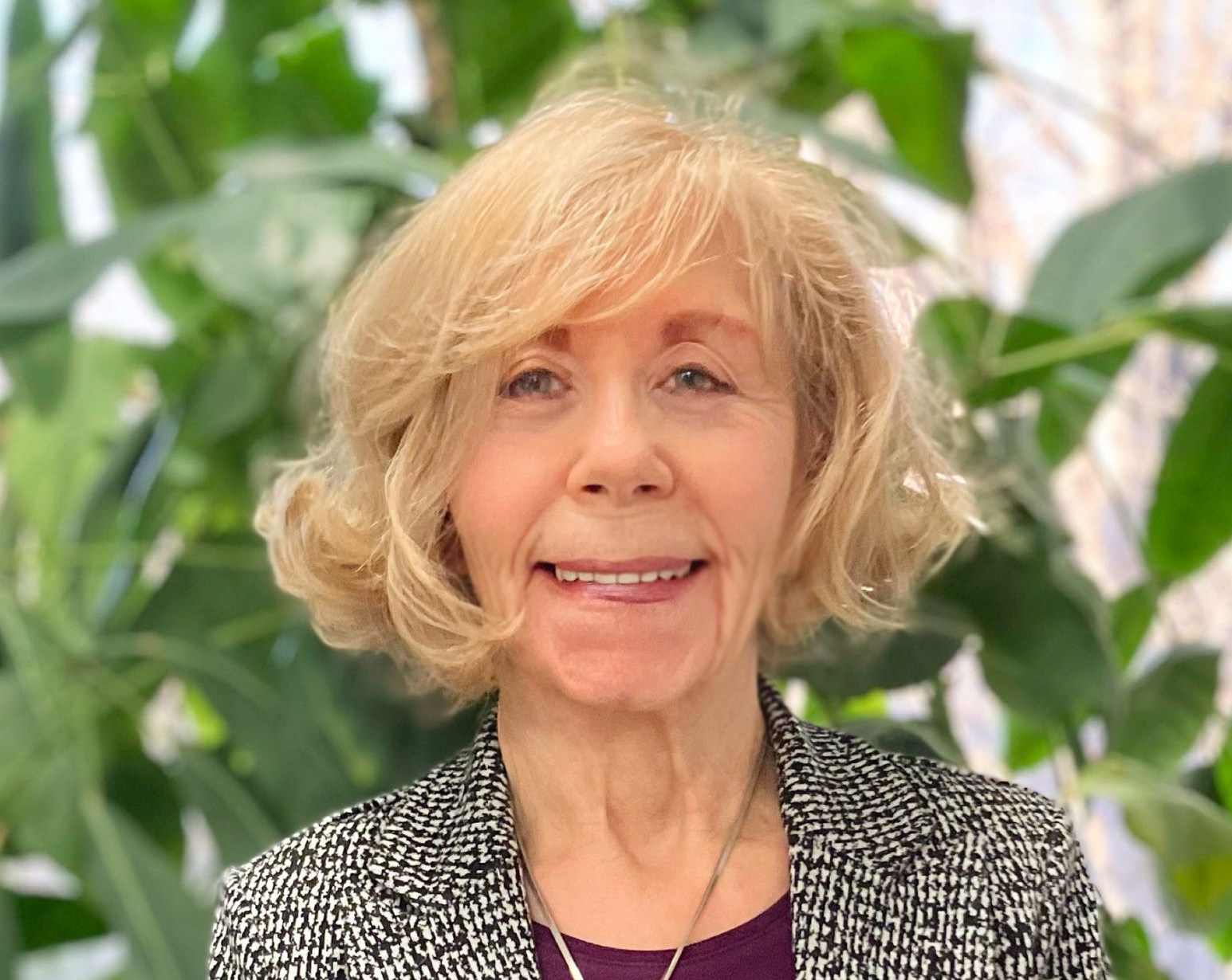 Woman with blonde hair wearing a patterned jacket and purple shirt smiles, plants in the background.