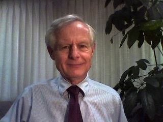 Man in blue striped shirt and purple tie, smiling, indoors near a plant and a white curtain.