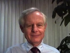 Man in blue striped shirt and purple tie, smiling, indoors near a plant and a white curtain.
