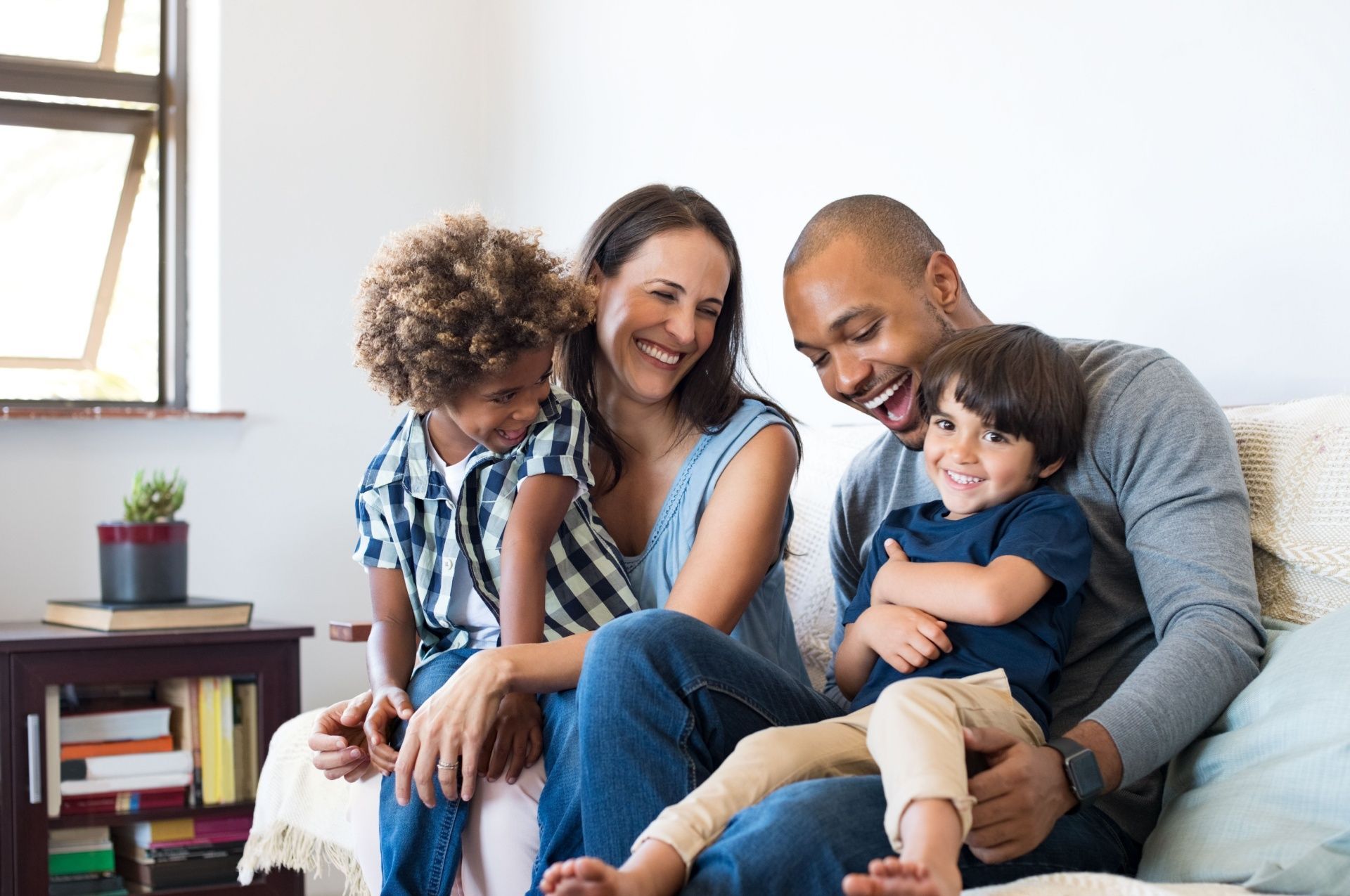 Family laughing together on a couch; two children and two adults. Bright, interior setting.