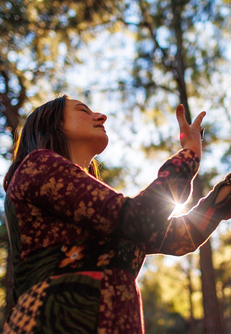 Woman reaching upwards in sunlight, hands open, forest background, warm tones.