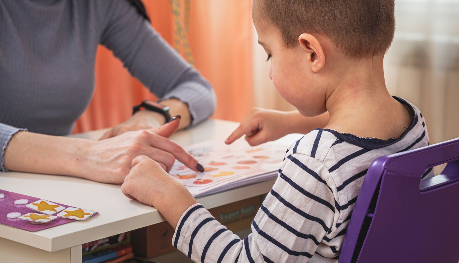 A child and adult point at a colorful worksheet on a table indoors.