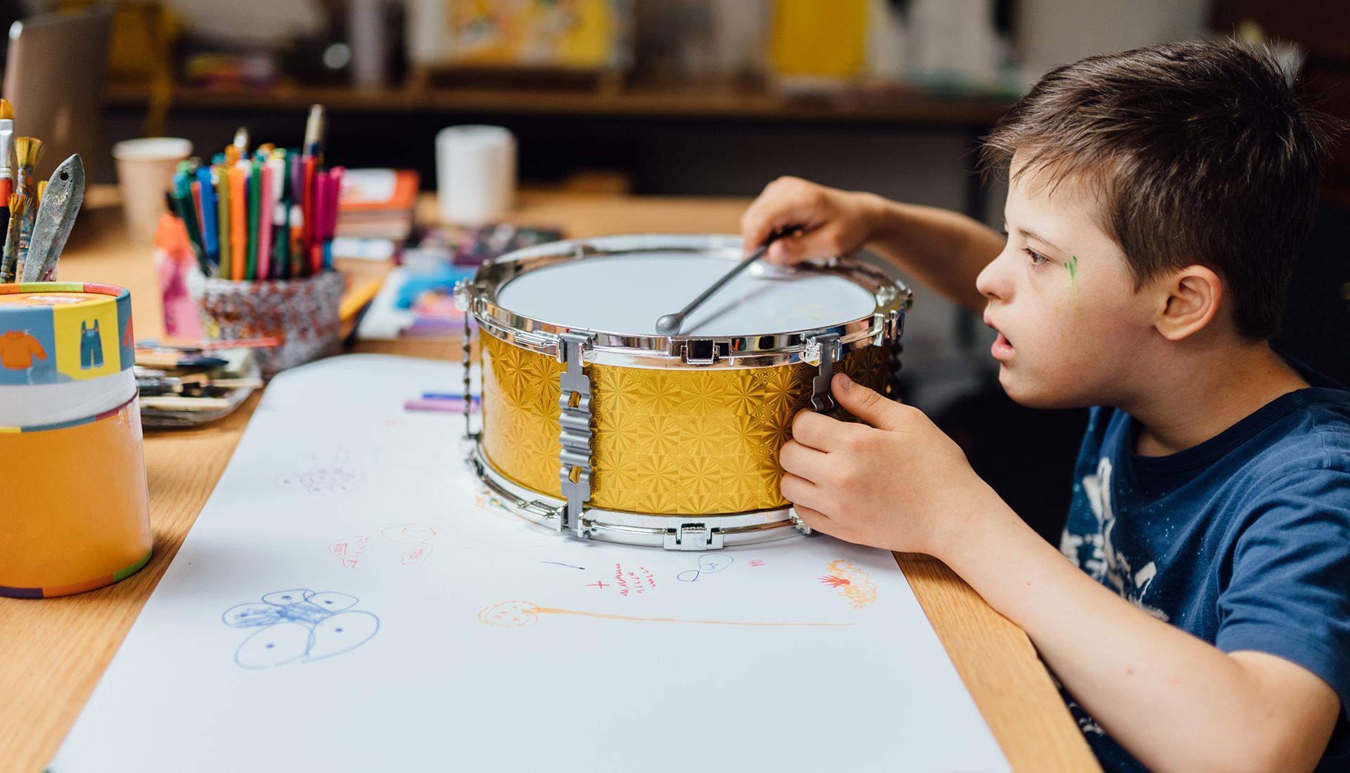 Boy with Down syndrome playing a drum on a table with drawing supplies.