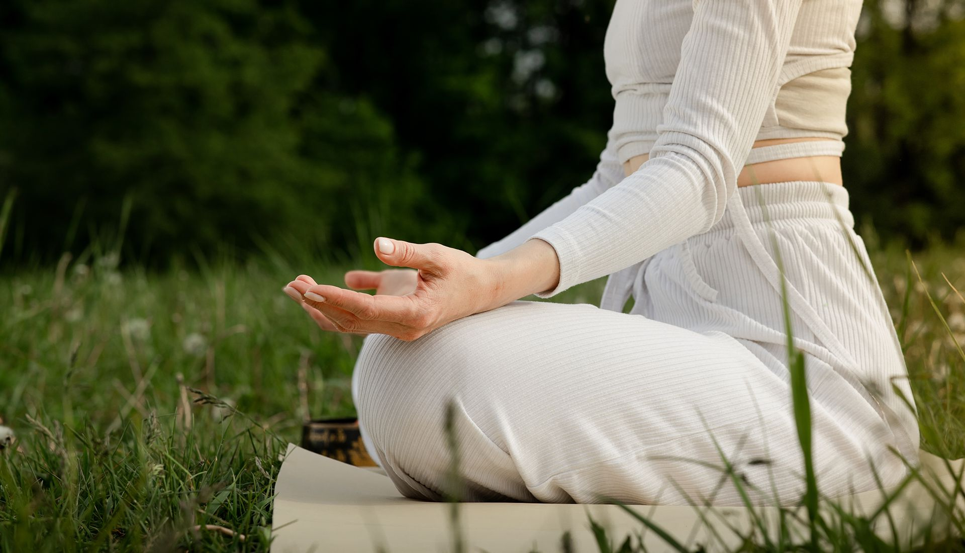 Person in white outfit meditating outdoors, hands in lap, grass and trees in background.